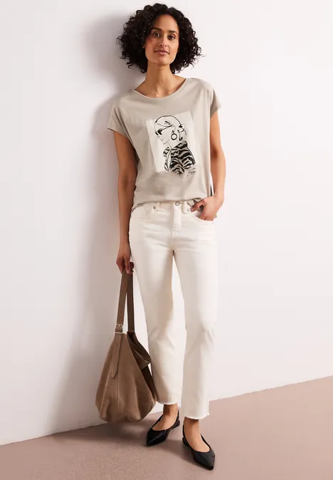 Frau in hellem Grafik-T-Shirt, weißer Hose, schwarzen Ballerinas und brauner Tasche vor neutralem Hintergrund.